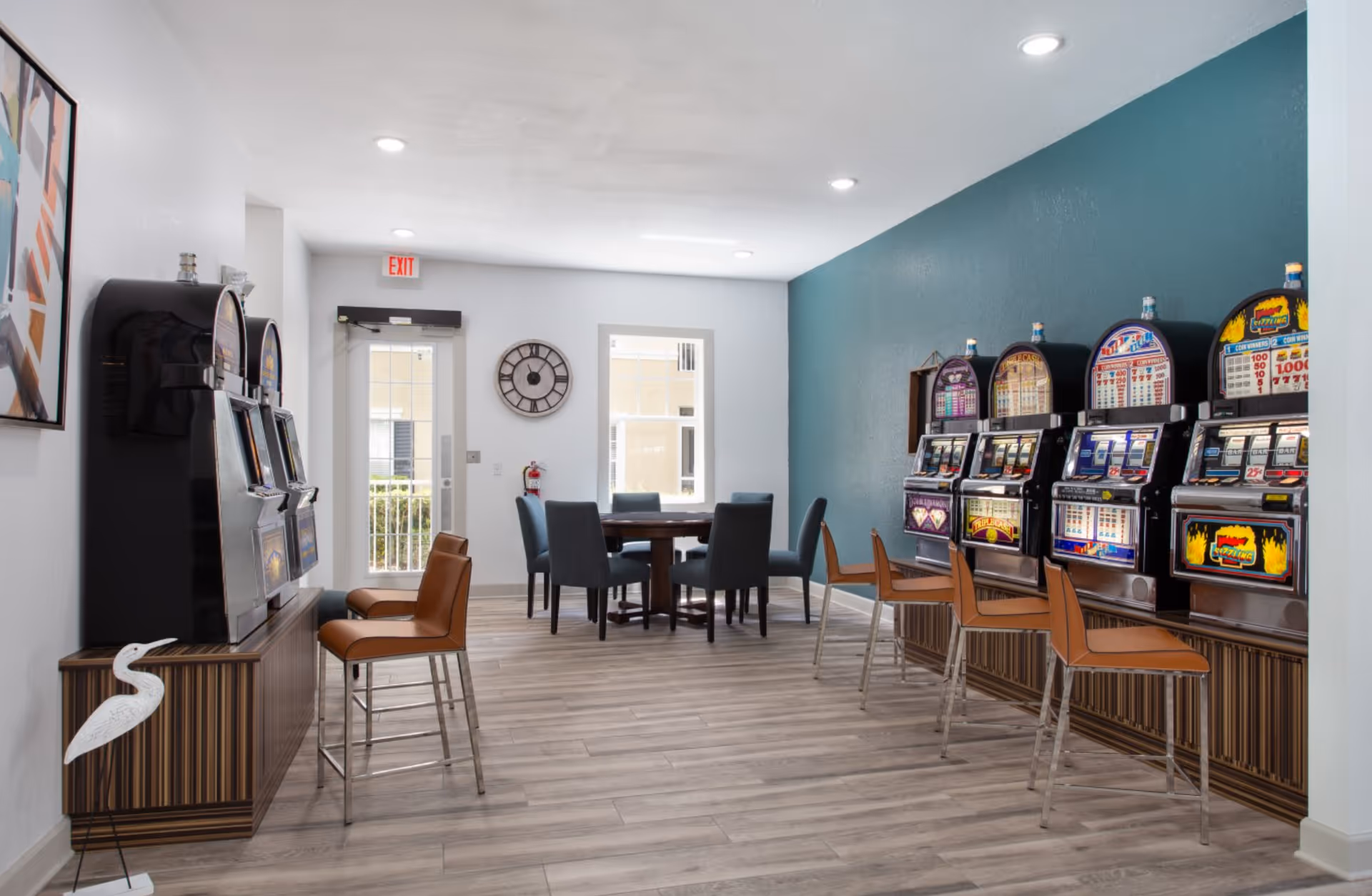 A bright room with several slot machines lined up against a teal accent wall, each accompanied by a brown chair. In the background, there is a round table with six blue upholstered chairs. The room has light wood flooring, white walls, a wall clock, and a glass door with an exit sign above it.