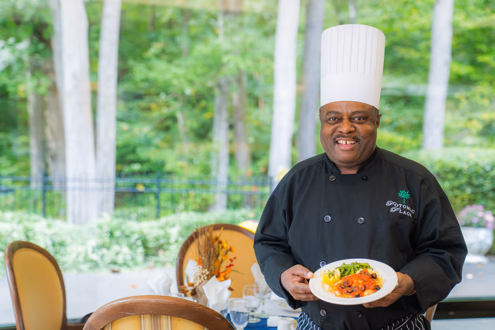 A smiling chef wearing a white chef hat and black uniform with the Potomac Place logo holds a plate of food in a dining area with large windows showing a green outdoor view.