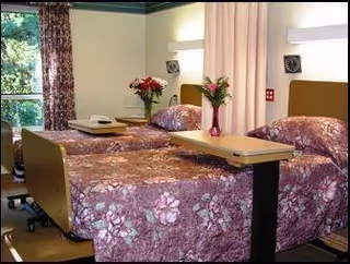 A senior living facility bedroom with two hospital-style beds covered in matching floral bedspreads. Each bed has an overbed table with a vase of flowers on it. The room has a window with floral curtains, light-colored walls, and wall-mounted lights above the beds.
