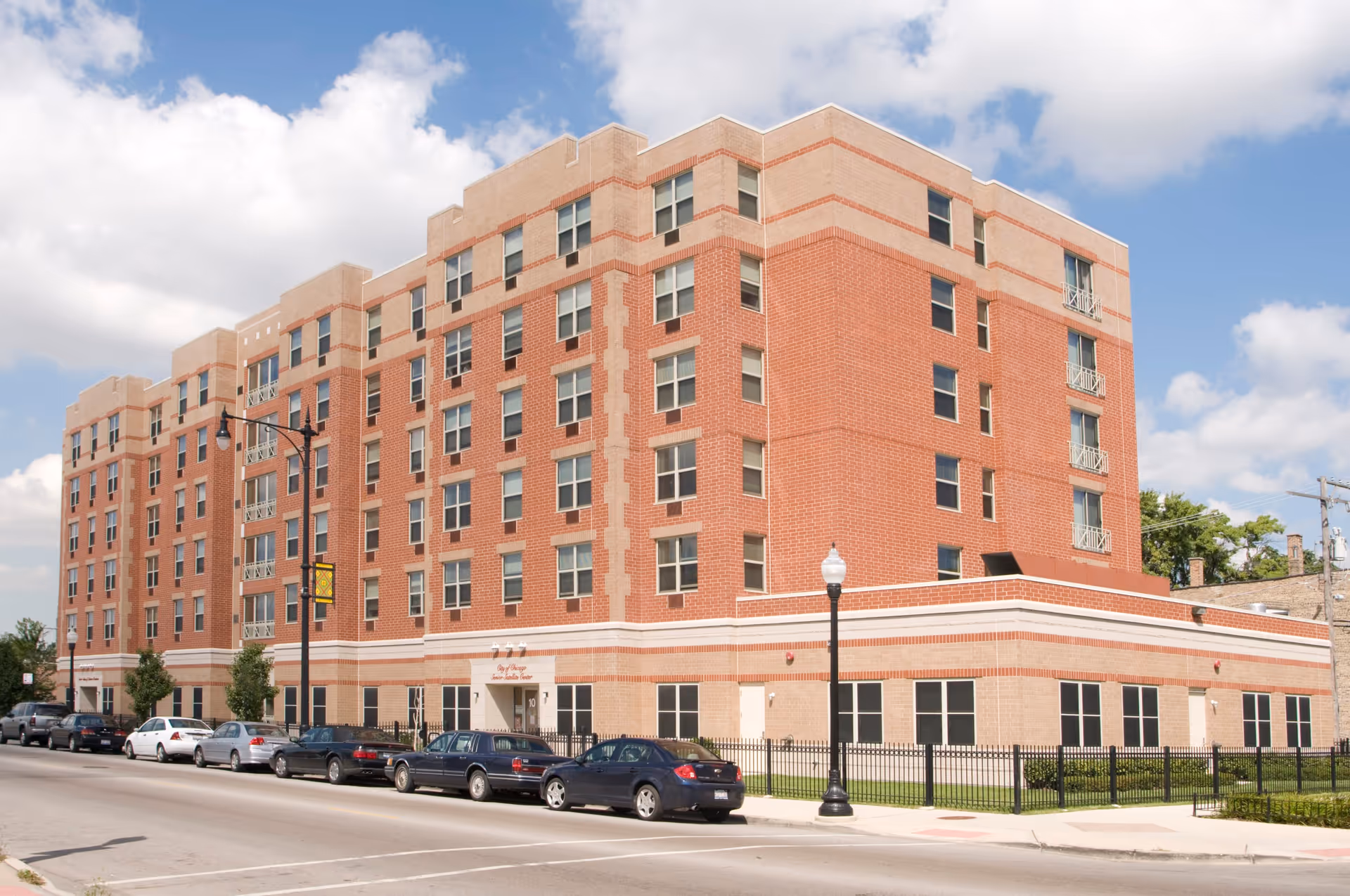 Exterior view of a multi-story brick building with several windows, a fenced area, parked cars along the street, and a clear sky with some clouds.