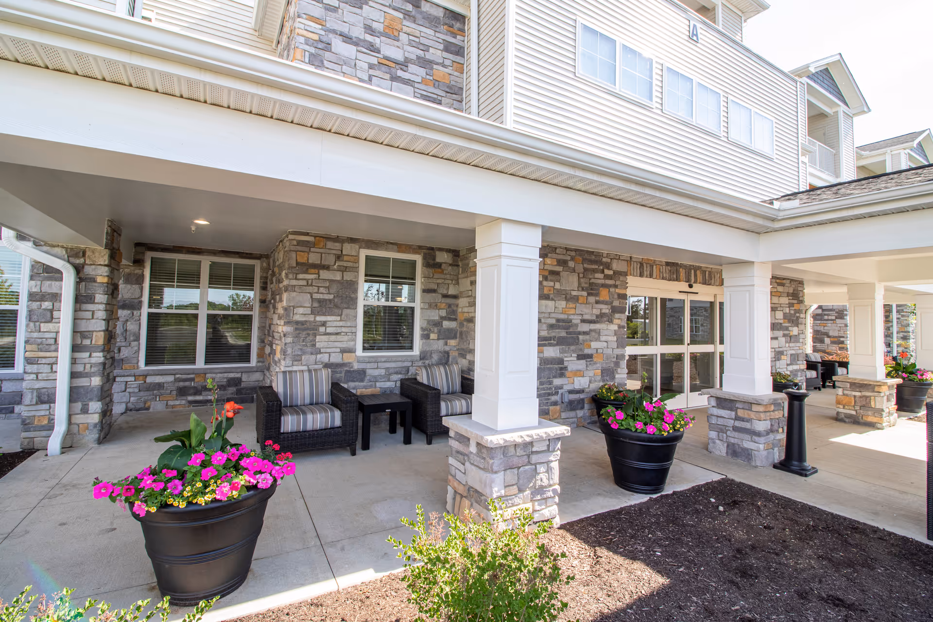 Covered outdoor seating area at StoryPoint Fairfield with stone pillars and walls, two black wicker chairs with striped cushions, a small black table, and large black planters filled with colorful flowers.