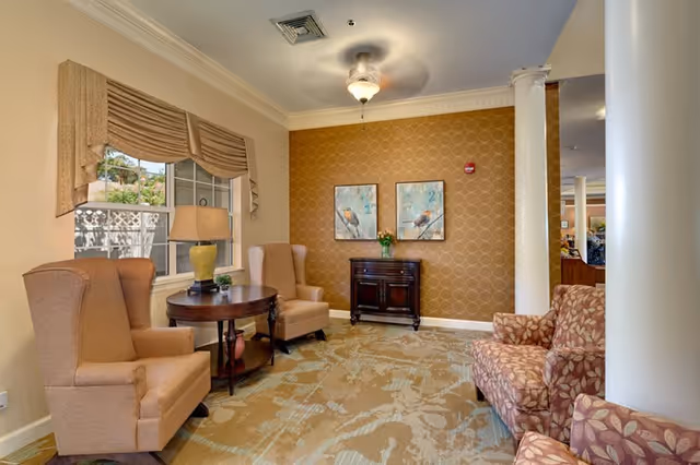 A cozy seating area with upholstered armchairs, a round side table and lamp, patterned wallpaper, and framed artwork.