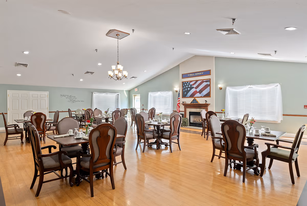 A spacious dining room with multiple wooden tables and chairs arranged neatly. Each table is set with white cups and small flower vases. The room has light green walls, large windows with white blinds, a chandelier hanging from the ceiling, and a fireplace with an American flag and a folded flag displayed above it.