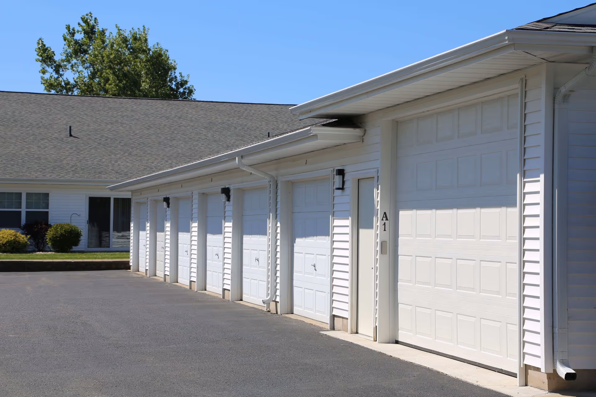 Several white garage doors along the exterior of a single-story building with a paved driveway and clear blue sky.