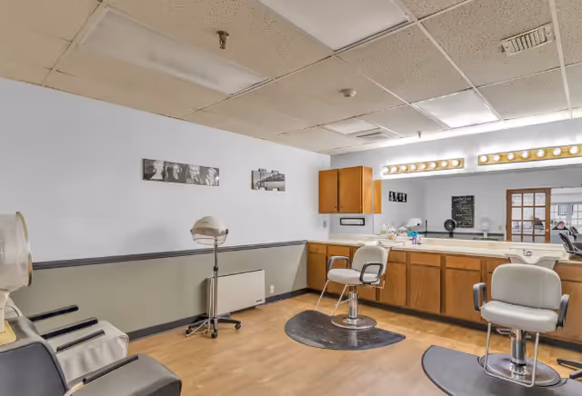 Interior view of a salon area in a nursing retirement home with two salon chairs on black mats, a hair dryer chair, wooden cabinets with a large mirror above, and framed black and white photos on the wall.