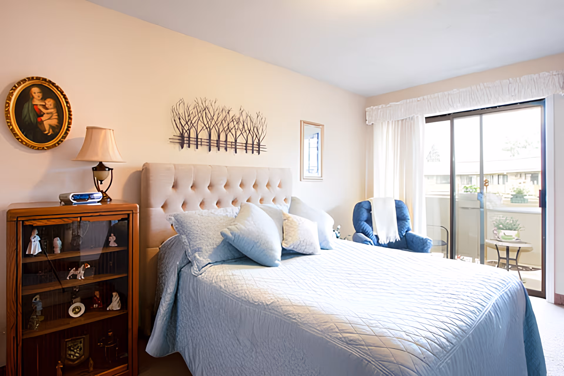 A bright bedroom with a large bed covered in a light blue quilt and multiple pillows. To the left of the bed is a wooden cabinet with glass doors displaying small figurines and a lamp on top. Above the bed is a metal wall art piece depicting trees and a small framed picture. To the right, there is a blue armchair with a white throw draped over it, next to a sliding glass door leading to a balcony with outdoor furniture and plants.