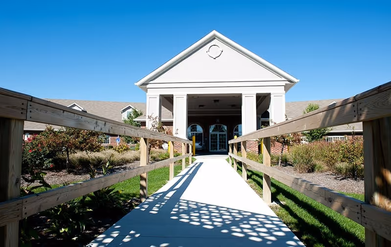 A paved walkway with wooden railings on both sides leading to the entrance of a single-story building with a white triangular pediment supported by columns. The building is surrounded by landscaped bushes and grass under a clear blue sky.