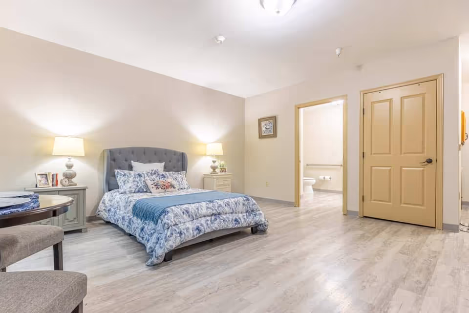 A spacious bedroom with a bed featuring a blue and white floral comforter and multiple pillows. There are two bedside tables with lamps and picture frames on each side of the bed. The room has light-colored walls and wood flooring. An open door reveals a bathroom with a toilet and grab bars.