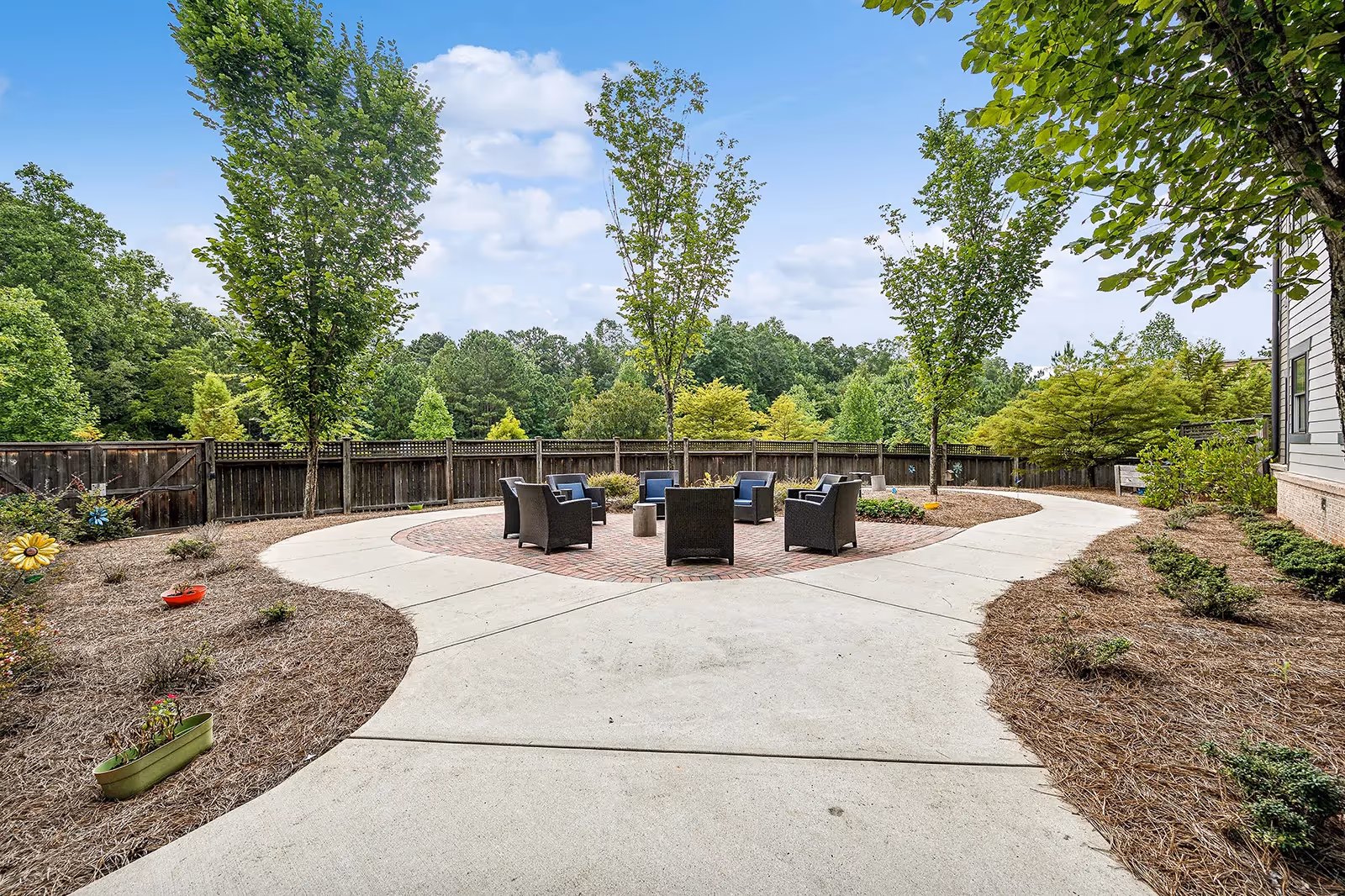 Paved outdoor courtyard with a circular brick seating area and chairs surrounded by trees and landscaped beds.