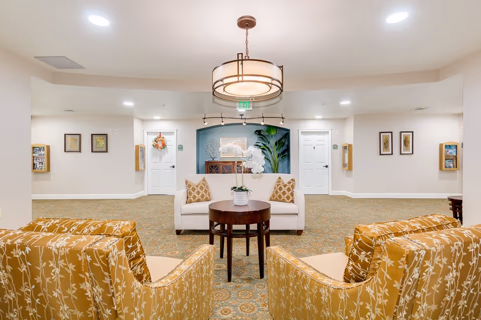 A well-lit common area with two patterned mustard yellow armchairs facing a white loveseat with two patterned pillows. A round wooden table with a white orchid plant is placed between the seating. The room has a patterned carpet, white walls with framed pictures, and two white doors in the background. A modern circular ceiling light fixture hangs above the seating area.