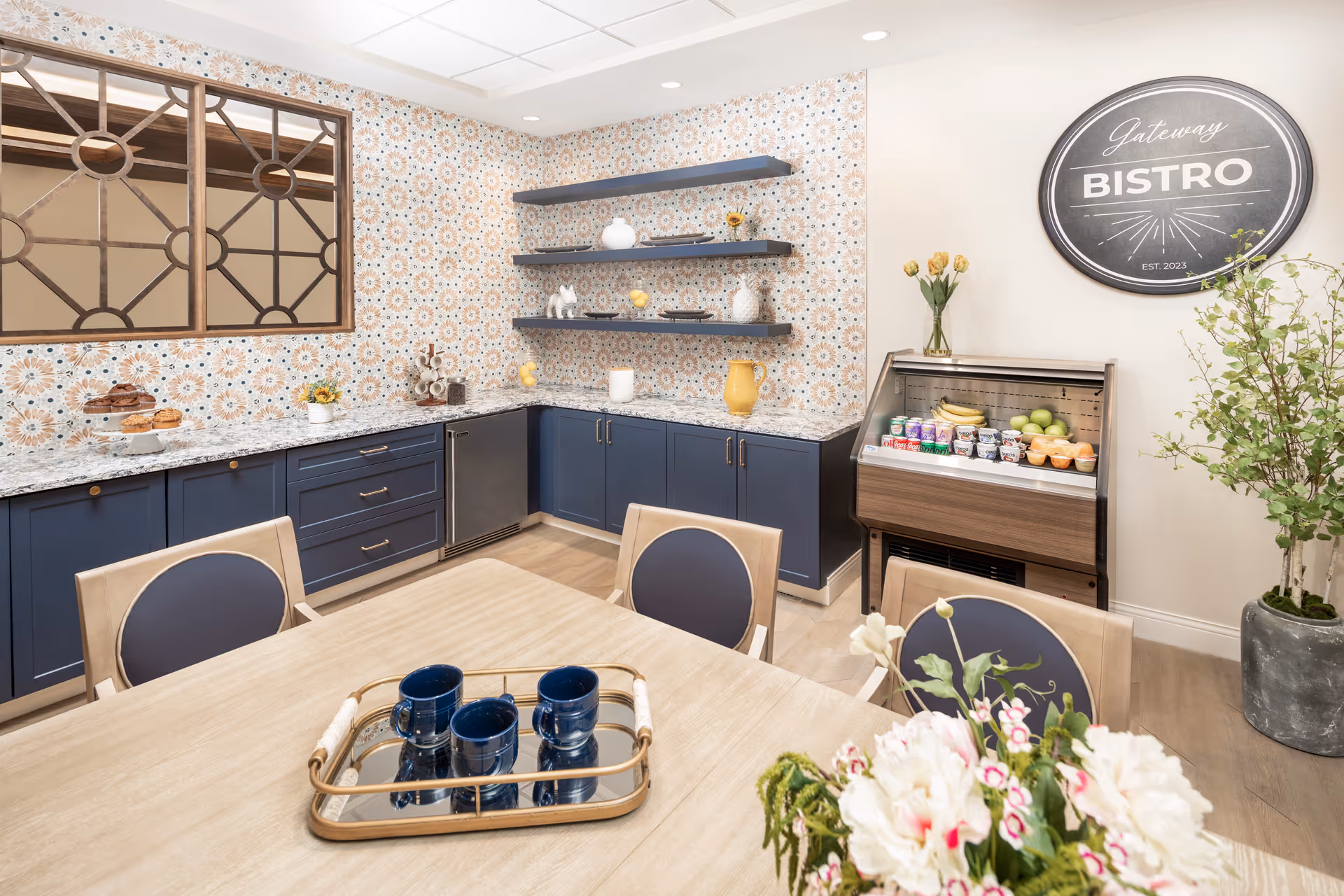 A bright and inviting bistro area with a wooden table and chairs in the foreground, a tray with blue mugs on the table, blue cabinets with a marble countertop along the wall, decorative patterned wallpaper, floating shelves with decorative items, a small refrigerated display case with fruit and yogurt, a vase with yellow flowers, and a round sign on the wall that reads 'Gateway Bistro EST. 2023'.