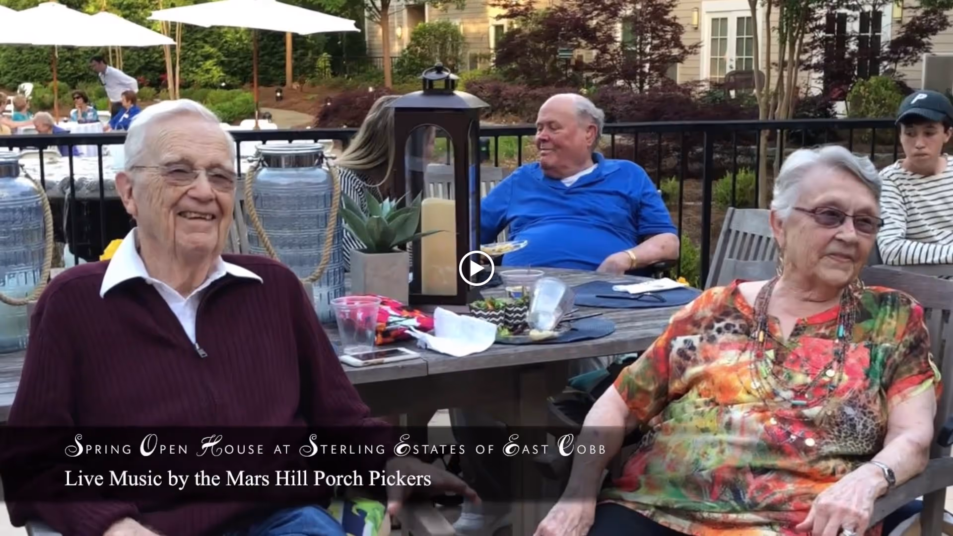 Two elderly people sitting outdoors at a wooden table with drinks and decorations, smiling and enjoying a social event. In the background, other people are seated under large white umbrellas in a garden or patio area.