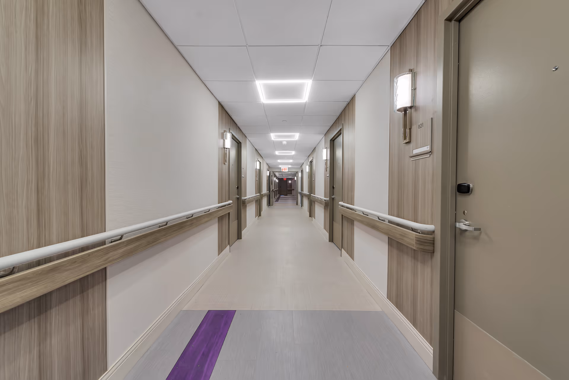 Long, well-lit corridor in an assisted living facility with handrails and resident room doors on both sides.