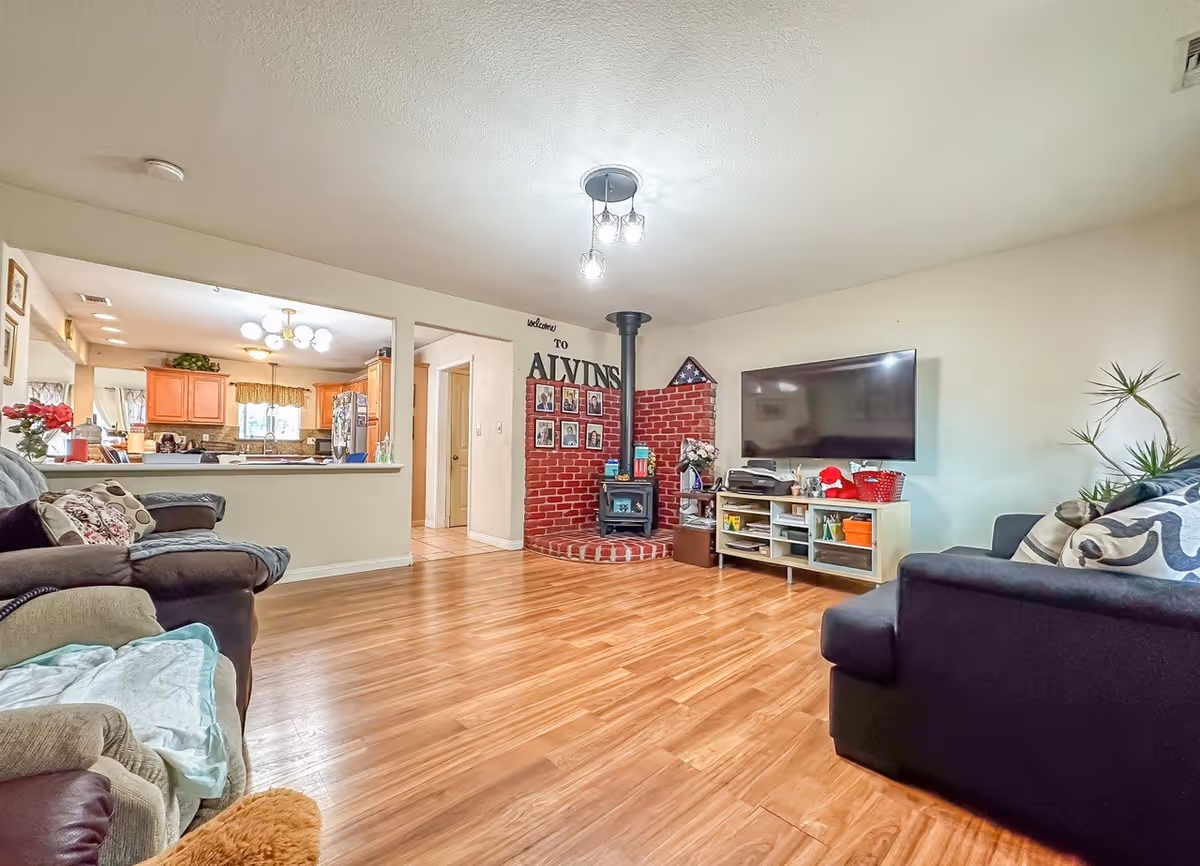 A cozy living room with wooden flooring, two sofas with cushions, a flat-screen TV mounted on the wall above a white entertainment unit, a small wood stove with a brick surround, and an open view into a kitchen with wooden cabinets and a window.