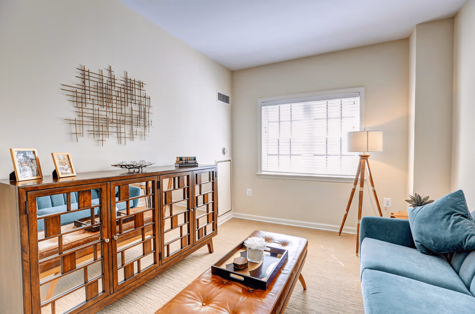 A bright and cozy living room with a blue sofa adorned with matching cushions, a wooden coffee table with a tray and decorative items, a wooden cabinet with mirrored doors, a modern floor lamp with a tripod base, and a large window with white blinds letting in natural light.