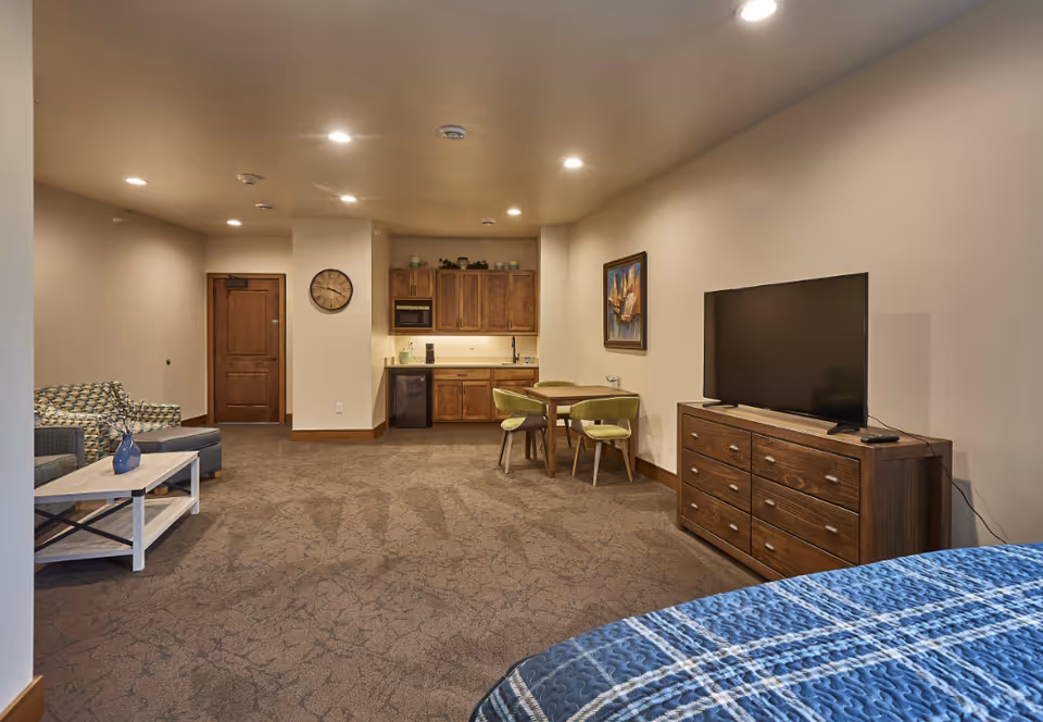 Interior view of a senior living facility room at TimberCrest at Glen Oaks featuring a bed with a blue patterned quilt in the foreground, a wooden dresser with a flat-screen TV on top, a small dining table with four green chairs, a kitchenette with wooden cabinets, a microwave, and a mini fridge, and a seating area with a patterned armchair and a coffee table with a blue vase. The room has beige walls, recessed ceiling lights, and a wall clock near the wooden door.