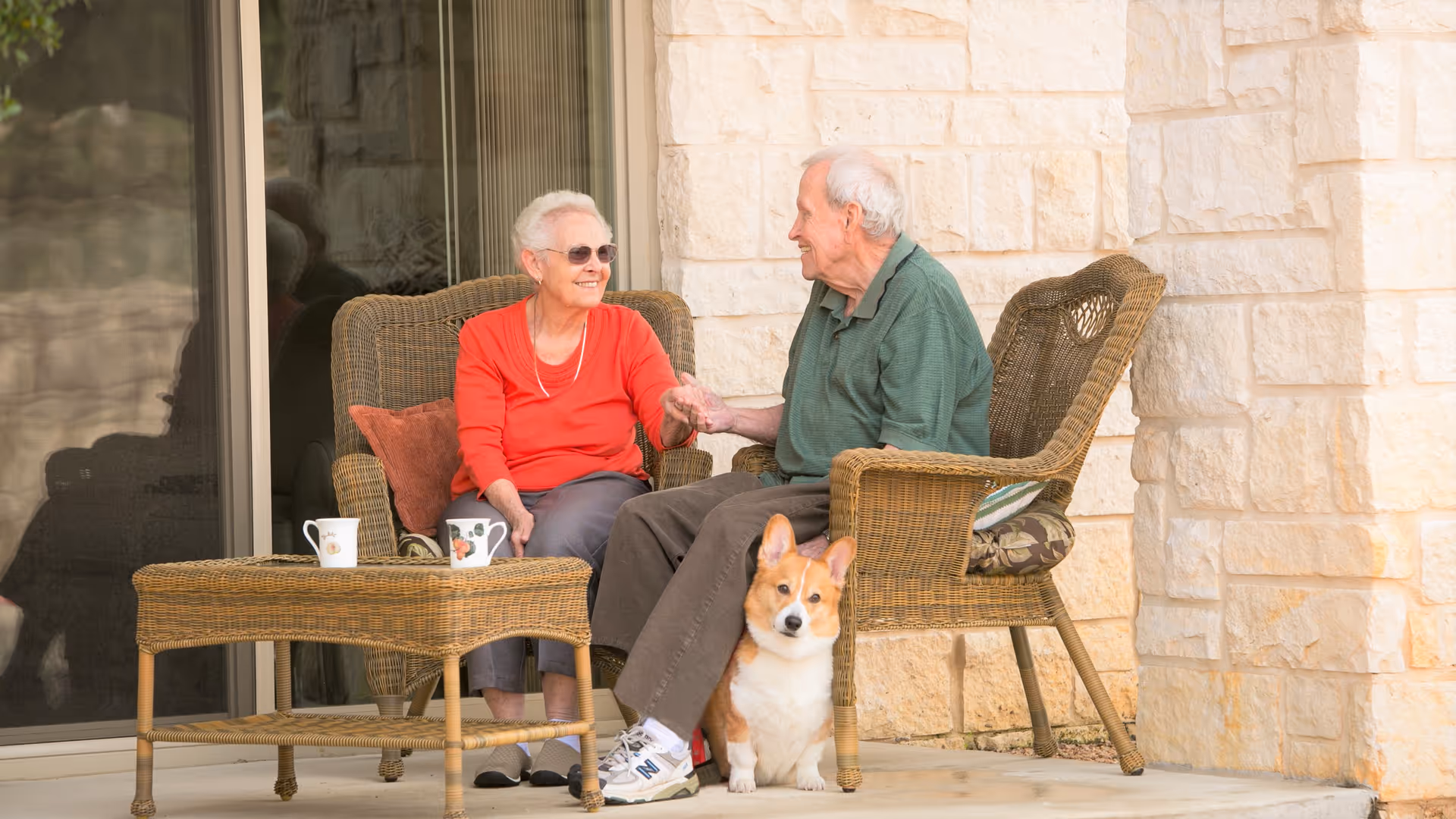An elderly couple sit on wicker chairs on a stone porch holding hands while a corgi sits at their feet.