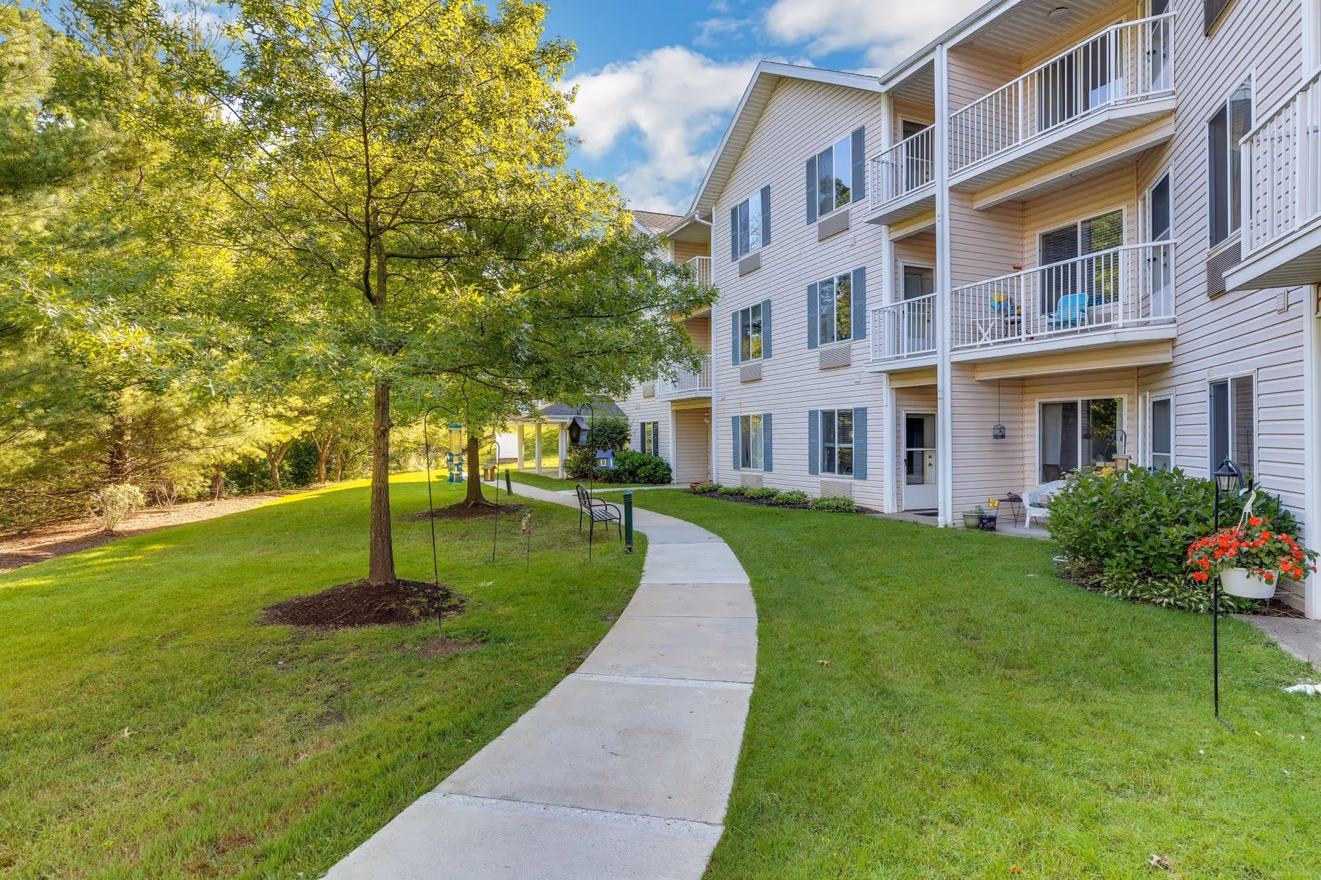 A paved walkway curves through a well-maintained grassy area with trees and benches, leading to a multi-story residential building with balconies and large windows under a partly cloudy sky.