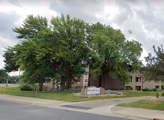 Brick three-story senior living building partially obscured by large trees with a stone entrance sign that reads 'Brookview'.