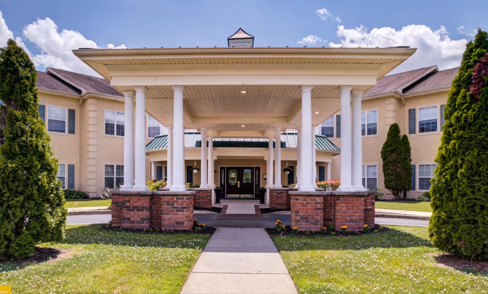 Front entrance of a senior living facility with a covered driveway supported by white columns and brick bases, surrounded by green grass, shrubs, and trees under a partly cloudy sky.