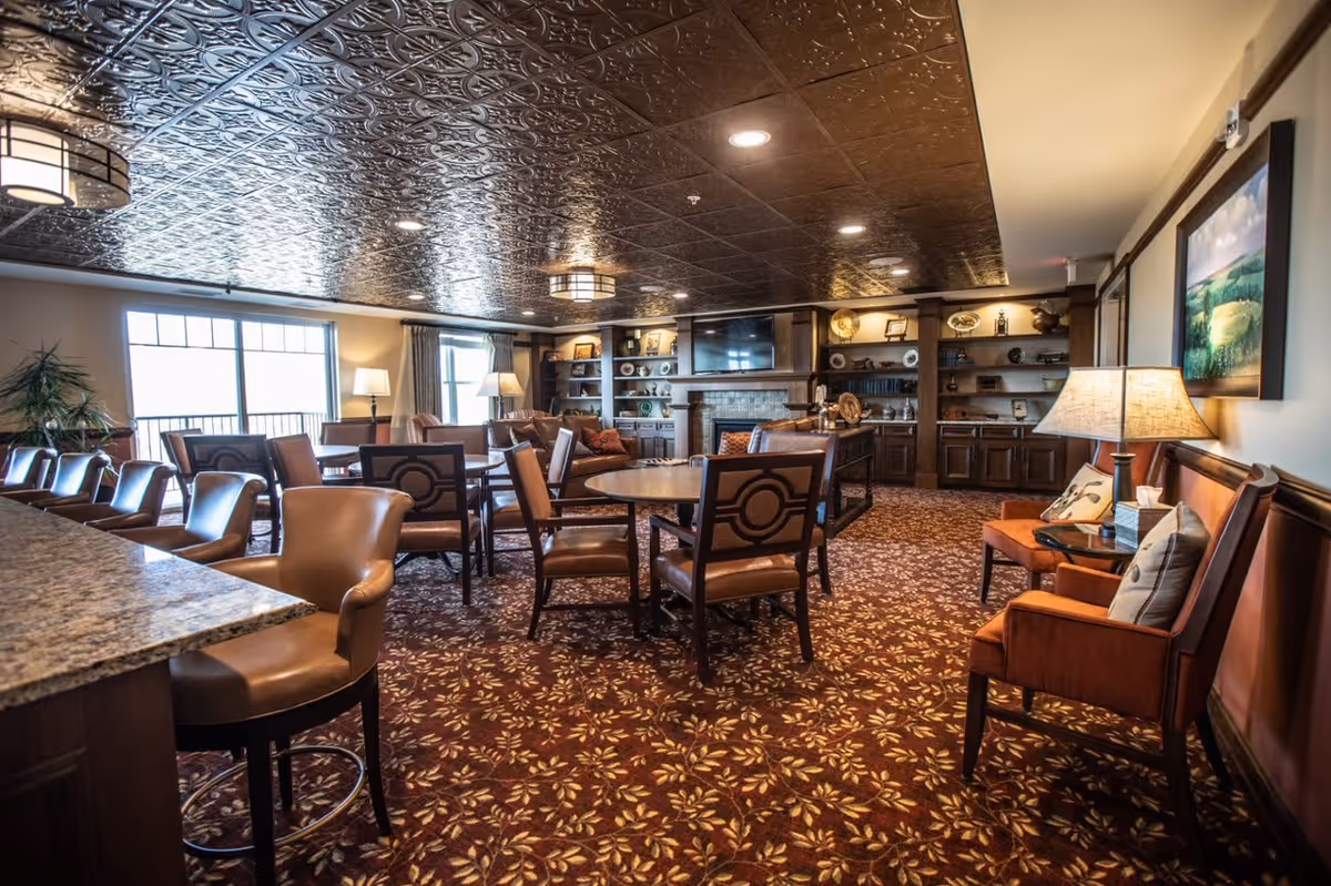 A spacious and warmly decorated common area with multiple round tables and chairs, leather armchairs, a patterned carpet, a decorative ceiling, built-in shelves with various decorative items, a fireplace, and a large window letting in natural light.