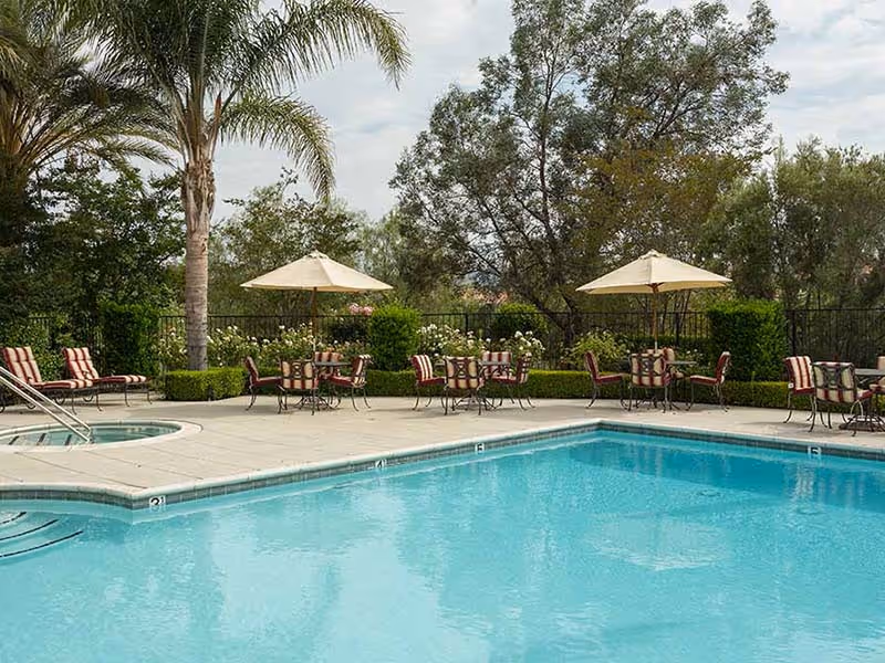 Outdoor swimming pool area with clear blue water, surrounded by a concrete deck. Several tables with umbrellas and chairs with red and white striped cushions are arranged around the pool. Palm trees and other greenery are visible in the background under a cloudy sky.