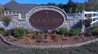 Stone sign with a large oval plaque reading 'The Courtyards Senior Living' surrounded by a landscaped garden bed with flowers and shrubs, with a white fence and building in the background under a clear blue sky.