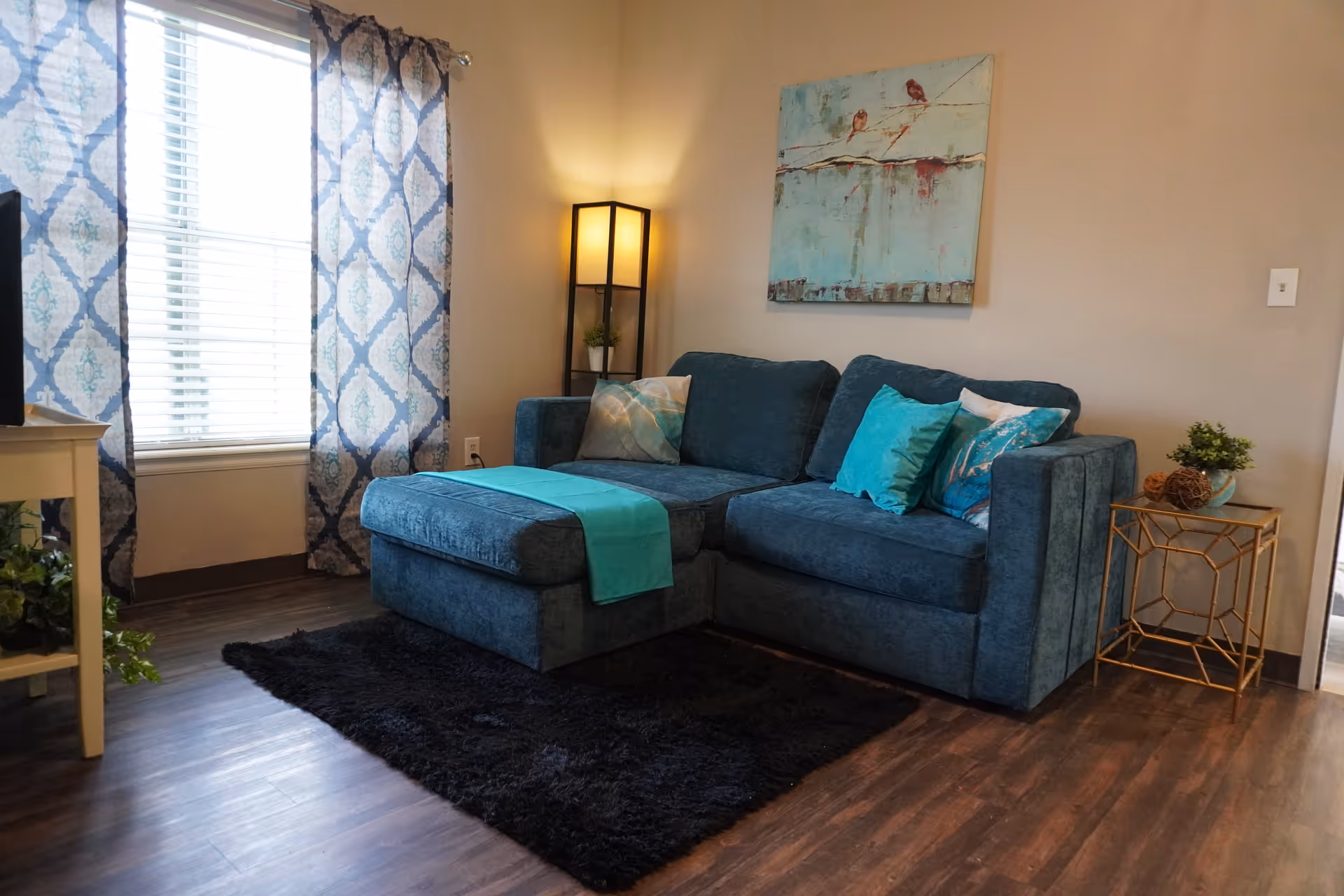 A cozy living room with a blue sectional sofa adorned with turquoise and patterned pillows. A turquoise throw is draped over the chaise section. The room features a black shag rug on wooden flooring, a tall floor lamp in the corner, a side table with decorative items, and a window with patterned blue and white curtains. A painting of birds on a branch hangs on the wall above the sofa.