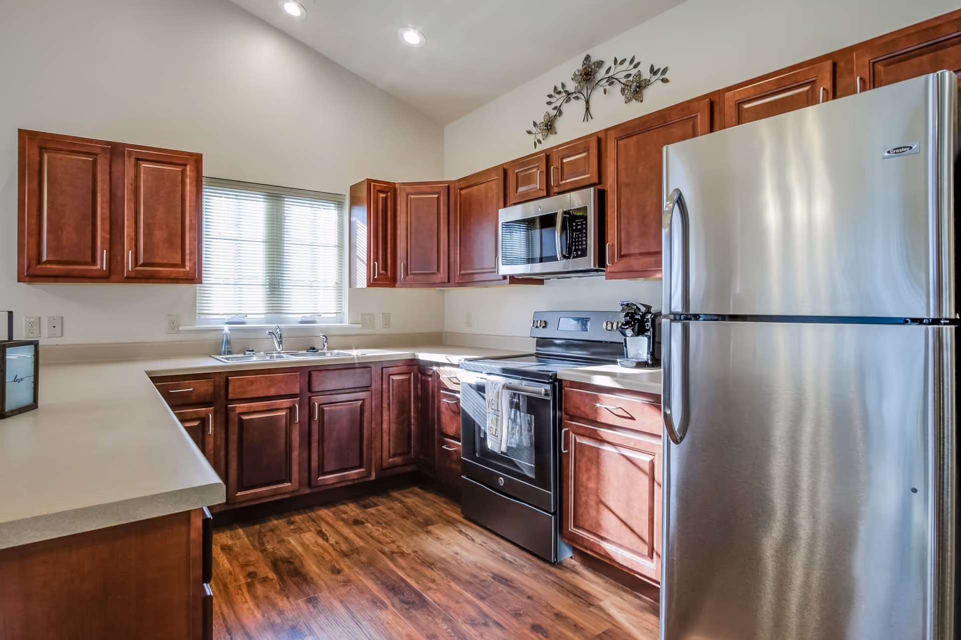 Bright kitchen with stainless steel refrigerator, stove and microwave, wooden cabinets, sink under a window, and wood flooring.