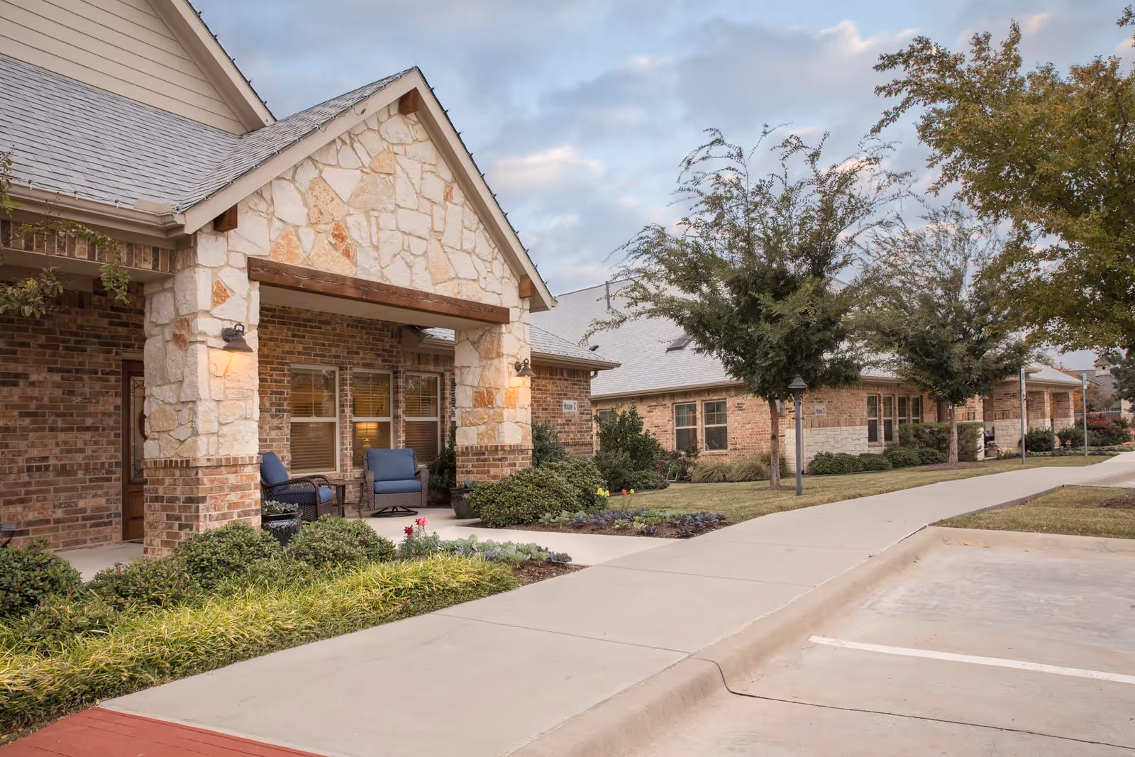 Exterior view of Mustang Creek Estates Frisco showing a stone and brick building with a covered porch area furnished with two blue chairs. The building is surrounded by landscaped greenery, trees, and a concrete sidewalk leading to a parking area.