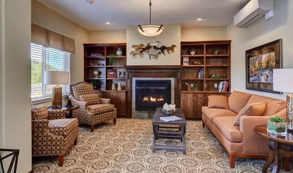 A cozy living room with a patterned carpet, two patterned armchairs, a peach-colored sofa, a wooden coffee table, and built-in wooden shelves flanking a fireplace with a metal horse sculpture above it. There is a window with blinds and a lamp on a side table, a framed picture on the wall, and an air conditioning unit mounted above.