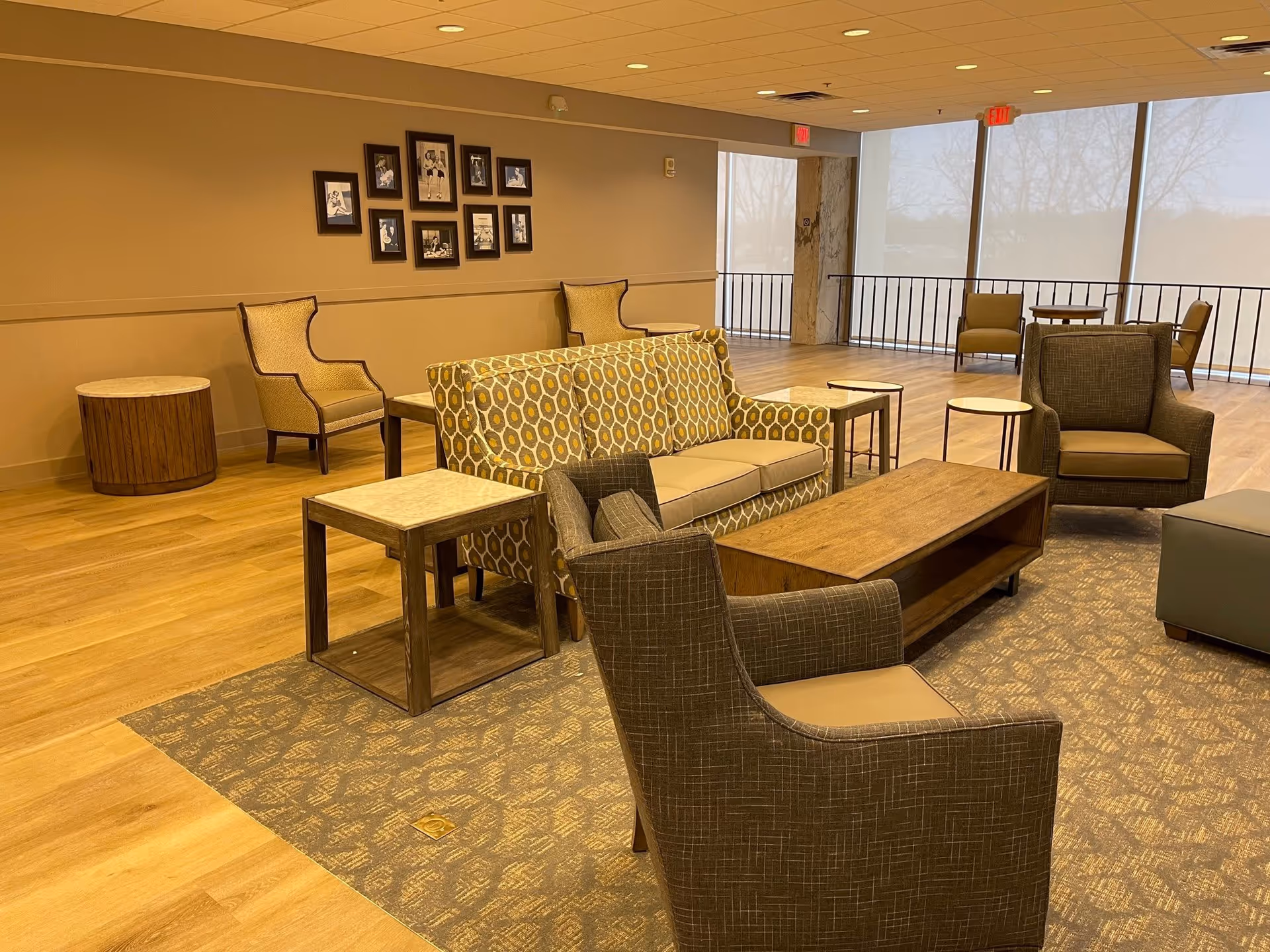 A spacious and well-lit common area in a retirement community featuring a patterned sofa, several armchairs, wooden coffee and side tables, a carpeted seating area, and large windows with roller shades allowing natural light to fill the room. Framed pictures are displayed on the wall.