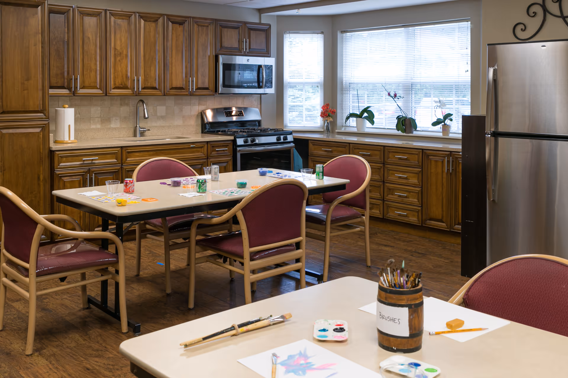 A kitchen area with wooden cabinets, a stainless steel refrigerator, stove, and microwave. There are two tables with chairs around them. One table has art supplies including paintbrushes, paint palettes, and paper with paintings. The other table has bingo cards, markers, and soda cans. The room has windows with blinds and some potted plants on the windowsill.
