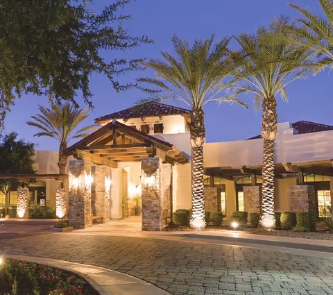 Exterior view of The Village at Ocotillo facility at dusk, featuring a well-lit entrance with stone pillars, palm trees, and a paved driveway.