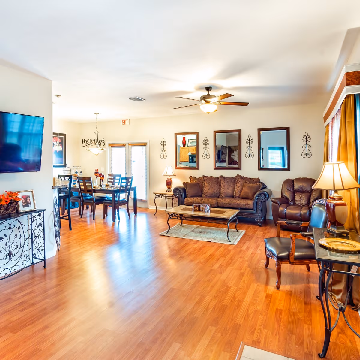 A bright and spacious living room and dining area with wooden flooring. The living room features a brown patterned sofa, a leather recliner, two chairs, a coffee table, and a side table with a lamp. Three mirrors and decorative wall hangings are on the wall behind the sofa. The dining area has a table set with four chairs and a hanging light fixture above. A flat-screen TV is mounted on the wall to the left, and there is a console table with framed photos and a plant. Large windows with curtains allow natural light to fill the room.