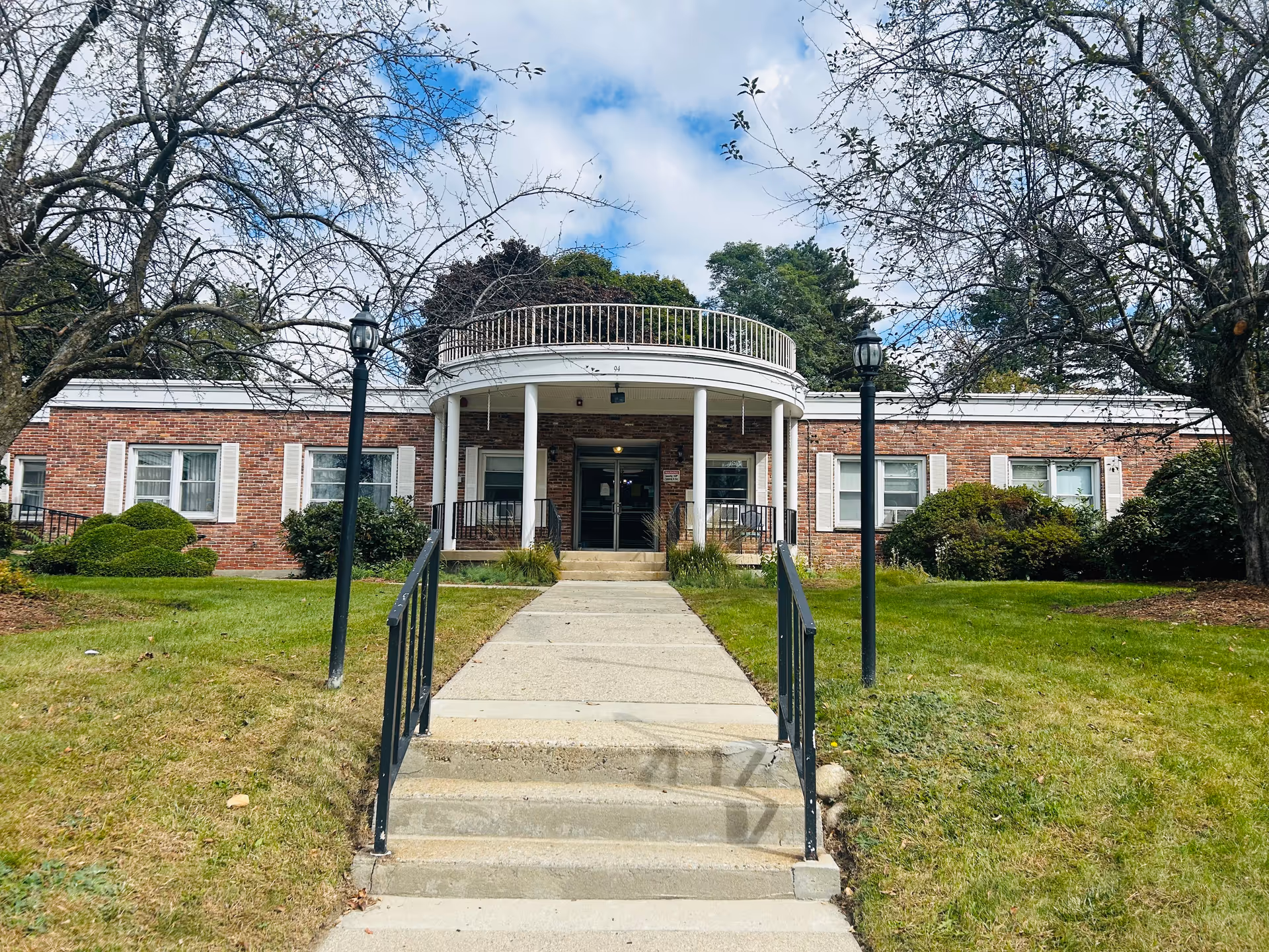 Front exterior view of a single-story brick building with white window shutters and a central entrance featuring a rounded portico supported by white columns. A concrete walkway with handrails leads up to the entrance, flanked by two black lamp posts. Trees and bushes surround the building under a partly cloudy sky.