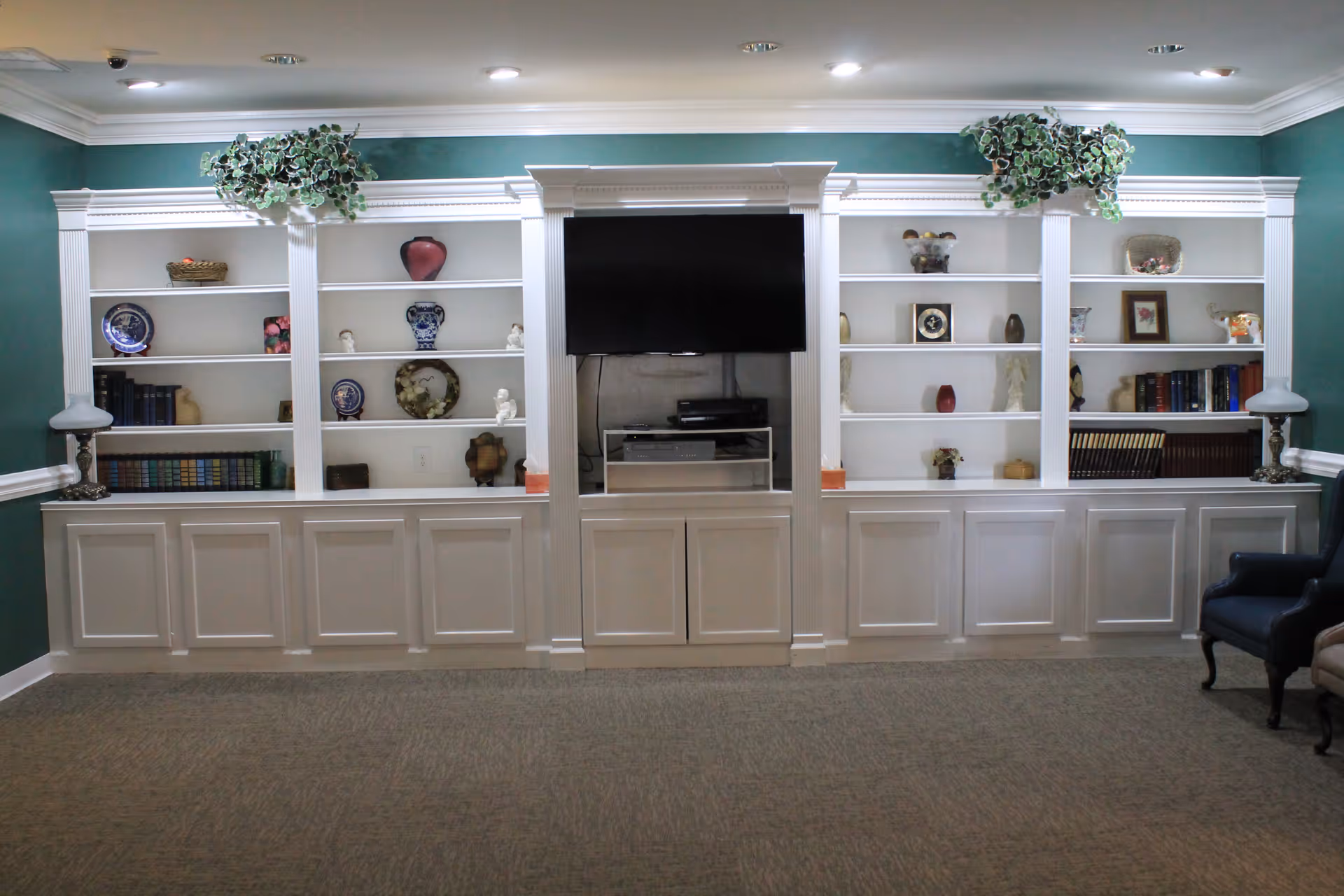 A living room area with a large white built-in shelving unit against a teal wall. The shelves contain decorative items such as vases, plates, books, and small sculptures. A flat-screen TV is mounted in the center of the shelving unit above a cabinet with electronic devices. Two lamps are placed on either end of the shelving unit, and there are two armchairs partially visible on the right side of the image. The ceiling has recessed lighting and white crown molding.