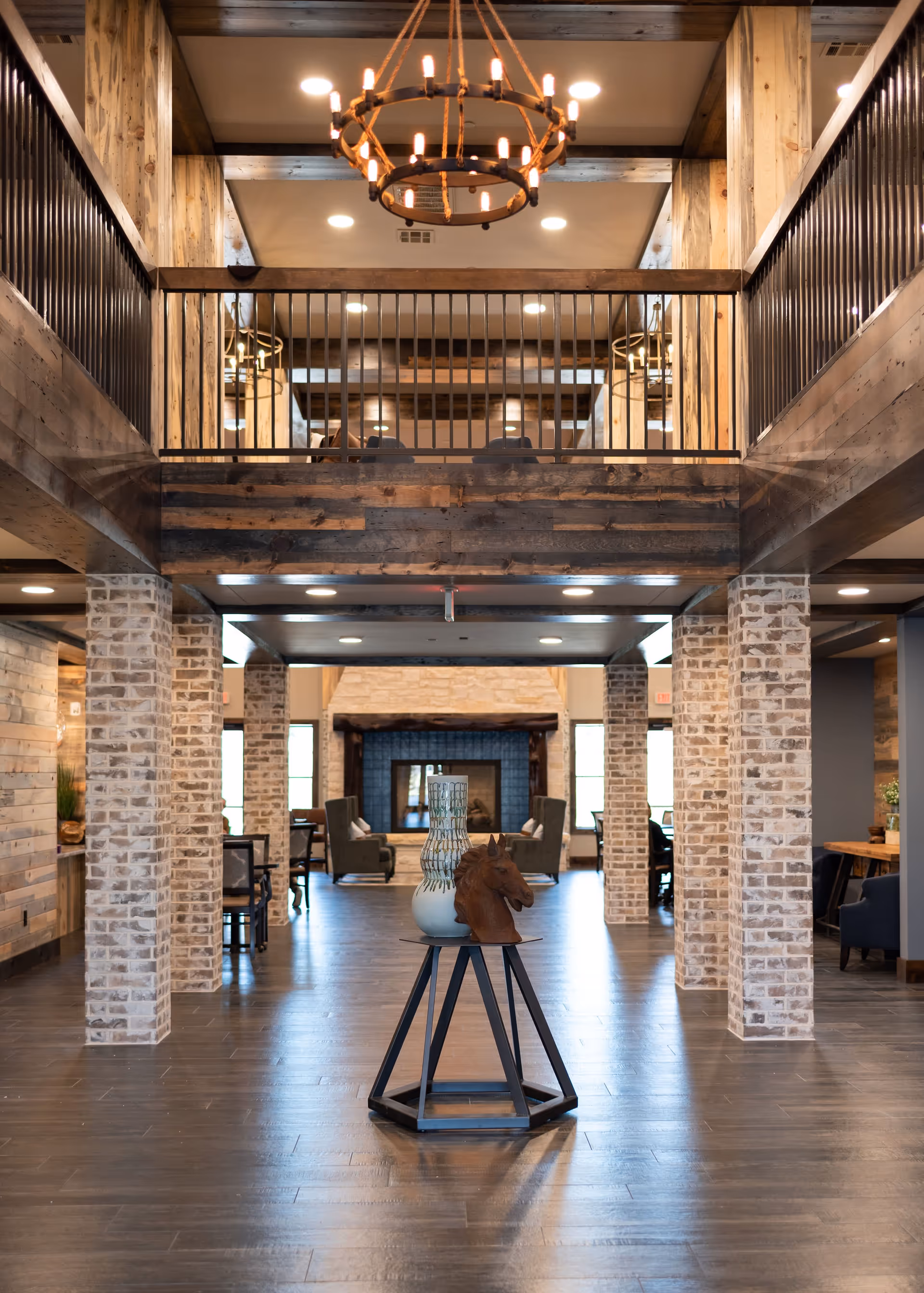 Spacious interior of Alders Rockwall featuring a rustic design with wooden beams, brick pillars, and a chandelier hanging from the ceiling. In the center, there is a small table with a decorative horse head sculpture and a vase. In the background, there are seating areas with armchairs arranged around a fireplace.