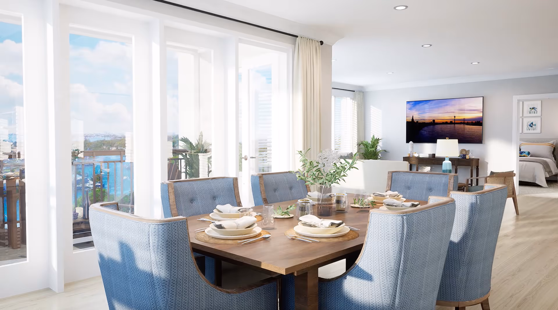 A bright dining area with a wooden table set for six, featuring blue upholstered chairs. The room has large glass doors leading to a balcony with a view of water and greenery. In the background, there is a living area with a TV mounted on the wall and a glimpse of a bedroom.