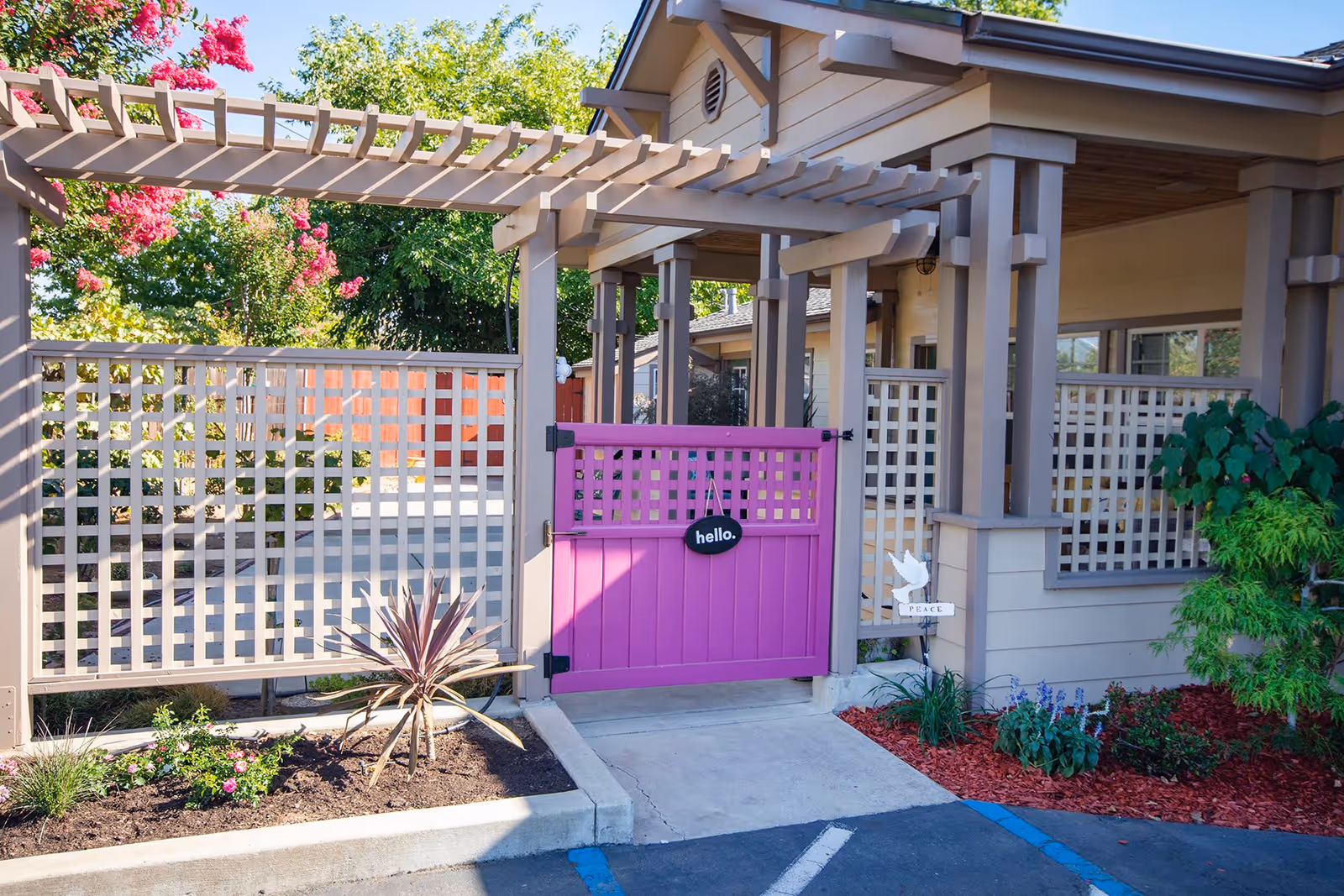 Entrance gate to a facility with a bright pink gate featuring a small sign that says 'hello.' The gate is part of a beige wooden fence with lattice design, surrounded by landscaped plants and flowers. The building behind has a covered porch area with columns and additional lattice fencing.