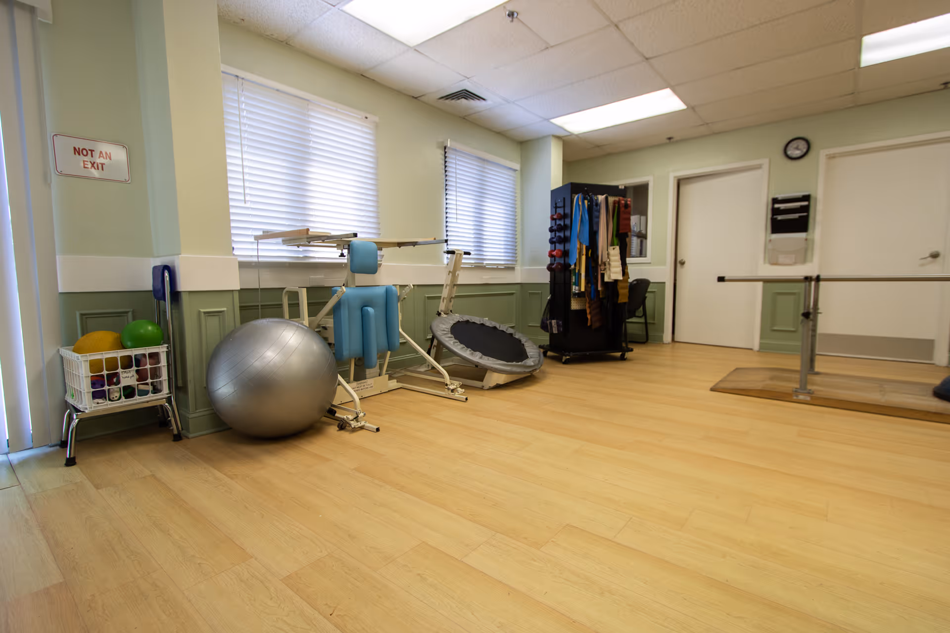 A rehabilitation exercise room with light wood flooring and pale green walls. The room contains various physical therapy equipment including a large silver exercise ball, a small trampoline, a blue padded exercise machine, a rack with resistance bands and weights, and parallel bars for walking exercises. Two windows with white blinds allow natural light into the room. A sign on the wall reads 'NOT AN EXIT'.