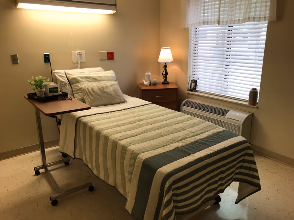 Small furnished single bedroom with a neatly made bed, overbed table, nightstand and a window with blinds.