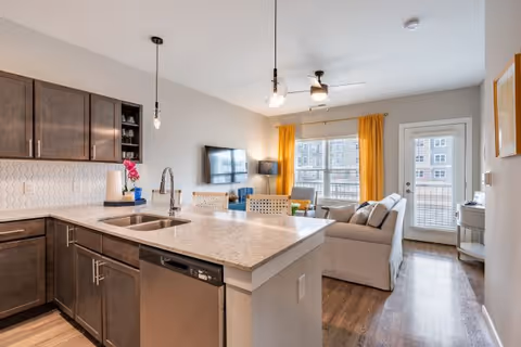 Open-plan kitchen and living area with a large island, stainless steel sink and appliances, a sofa facing a wall-mounted TV, and yellow curtains at the window.