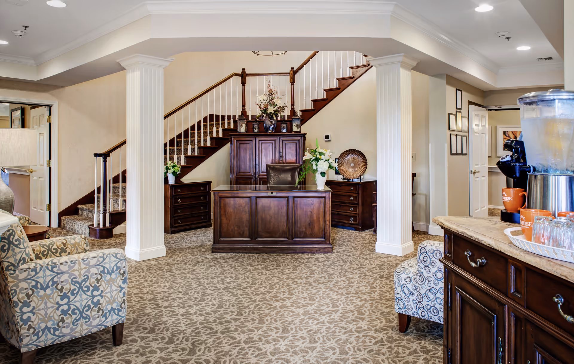 A spacious and elegant interior area featuring a wooden reception desk with a leather chair, flanked by two wooden cabinets with floral arrangements. Behind the desk is a staircase with a wooden banister and carpeted steps. The room has patterned carpet flooring, two white columns, and comfortable patterned armchairs. On the right side, there is a countertop with a beverage dispenser, coffee machine, orange mugs, and glasses.