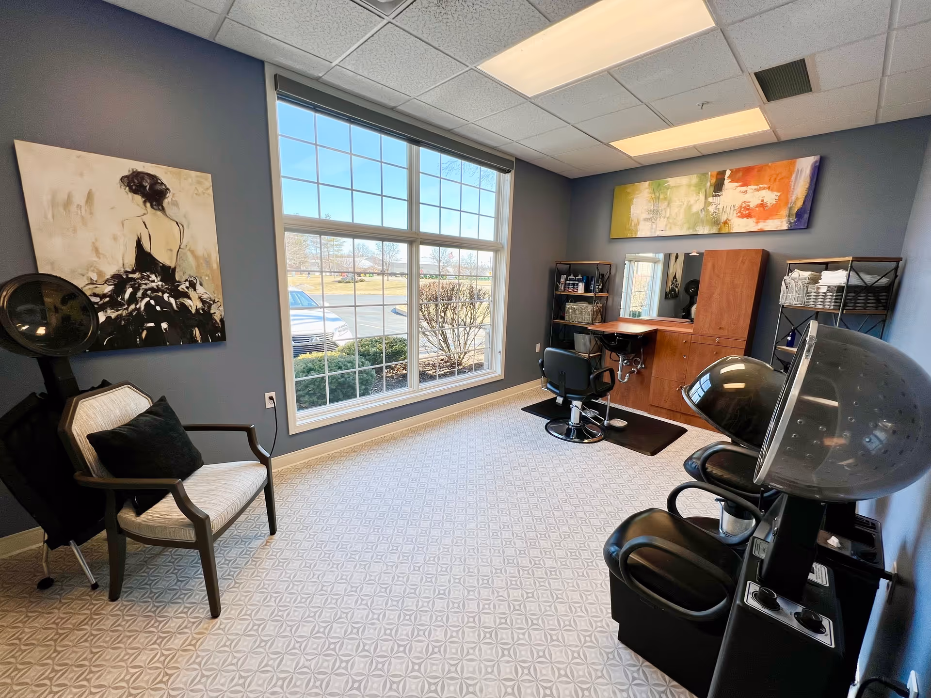 A bright and clean hair salon room with a large window showing an outdoor parking lot. The room has a salon chair in front of a wooden cabinet with a mirror, shelves with hair products and towels, a hair dryer chair, and a cushioned armchair with a black pillow. The walls are painted blue and decorated with two paintings, one of a woman in a black dress and an abstract colorful piece.