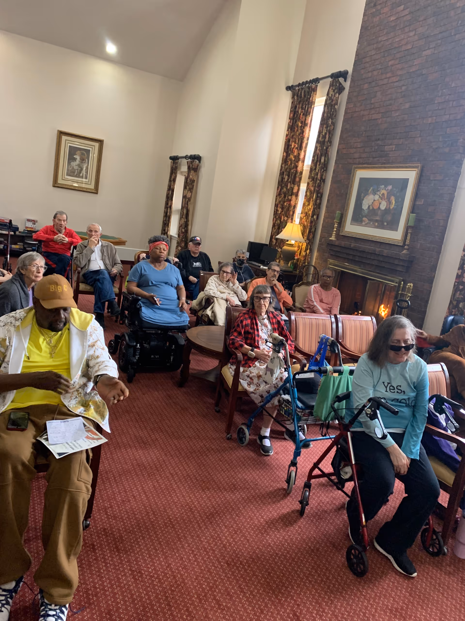 A group of older adults seated in a carpeted common room with high ceilings, a brick fireplace, and walkers.