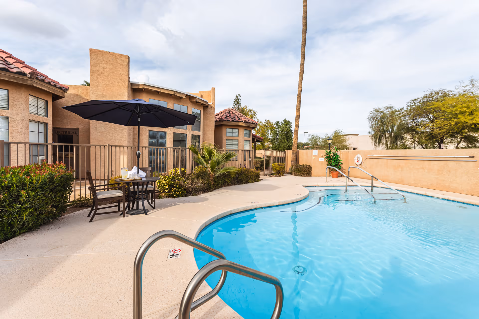 Outdoor swimming pool area at a senior living facility with clear blue water, metal handrails, a table with two chairs under a large black umbrella, surrounded by a beige building with windows and some greenery including bushes and a palm tree.