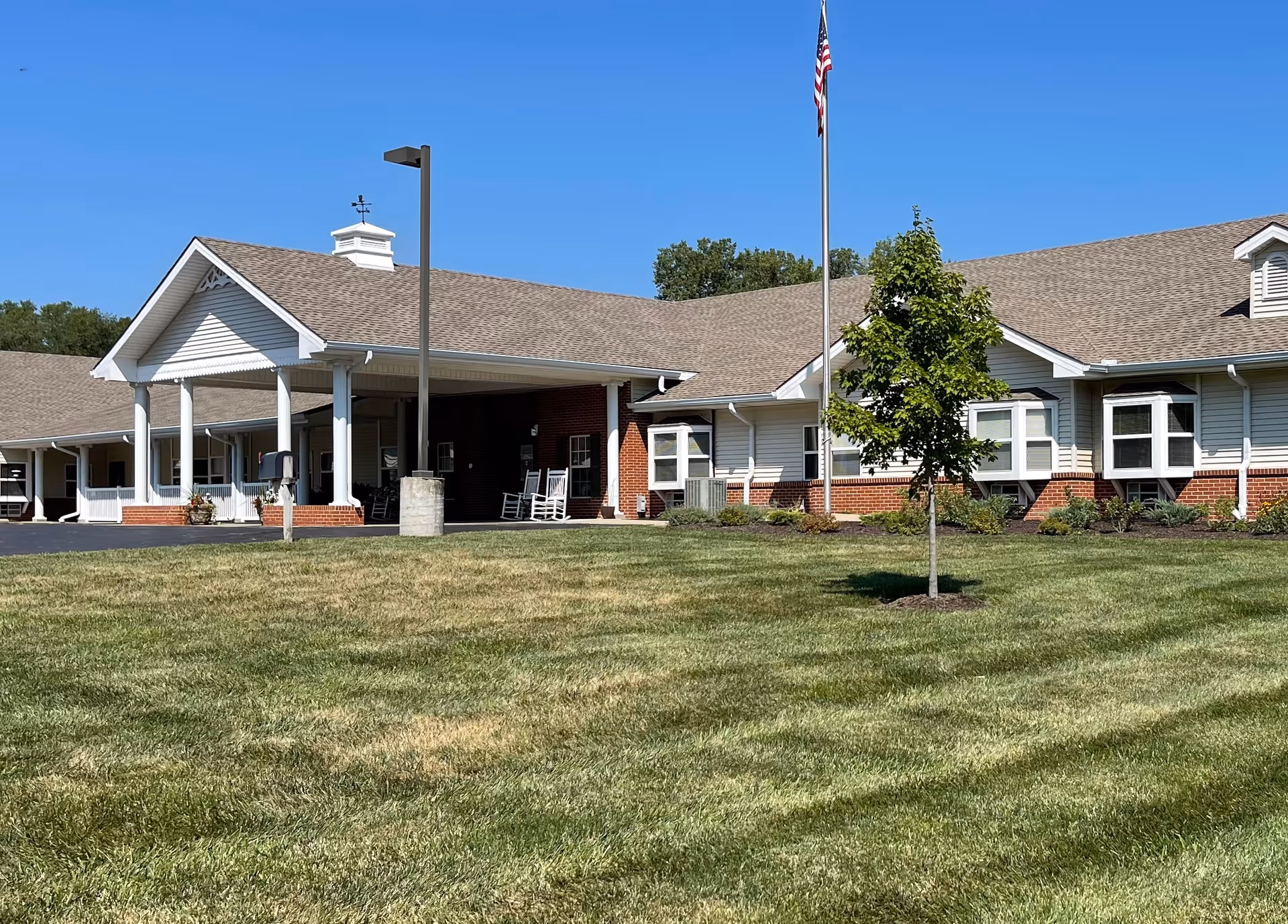 Exterior view of a single-story senior living facility building with a covered entrance supported by white columns, a flagpole with an American flag, a small tree, and a well-maintained lawn under a clear blue sky.