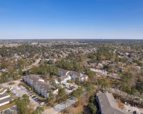 Aerial view of a residential area featuring multiple buildings surrounded by trees and parking lots under a clear blue sky.