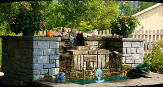 Outdoor stone fountain with cascading water, planters, and greenery in a courtyard.